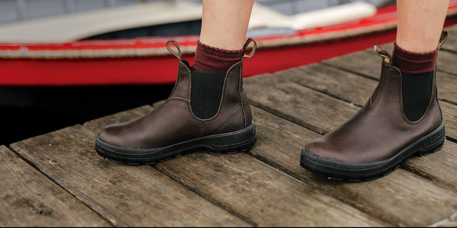 A person wearing dark brown Chelsea-style ankle boots with black elastic side panels, standing on a weathered wooden dock with boats blurred in the background.