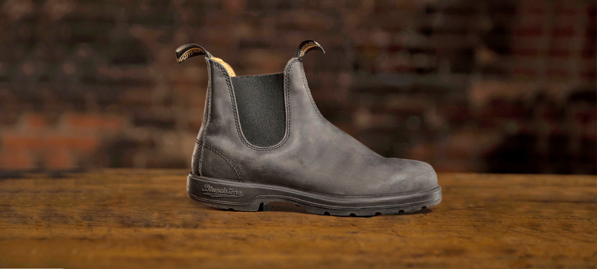Rustic Black Chelsea boot on a wooden surface with a brick wall background